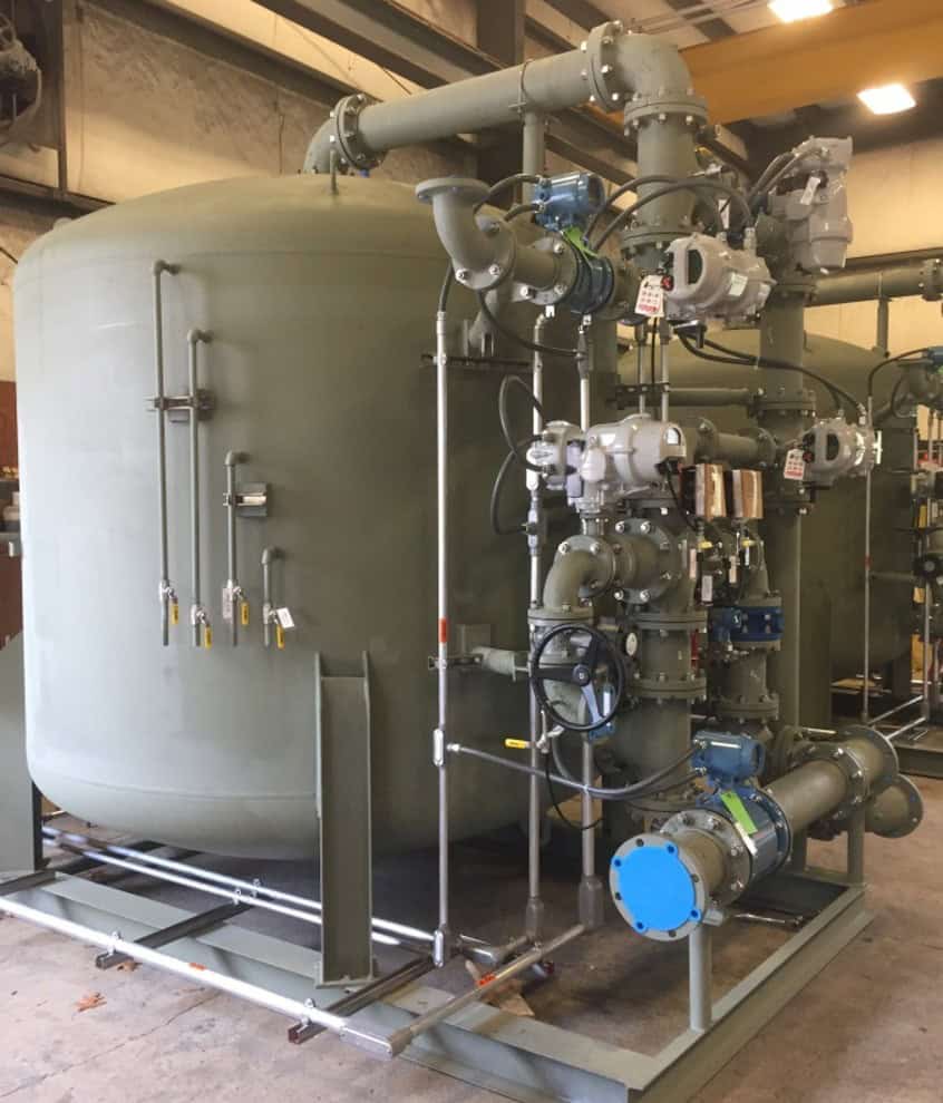 Large olive-green industrial pressure vessel with pipes, valves, gauges, and a blue pump mounted on a metal skid.