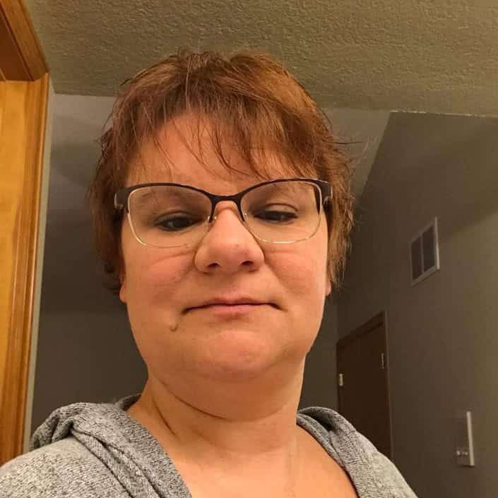 Person with short reddish-brown hair and glasses wearing a gray hoodie, taking a selfie indoors